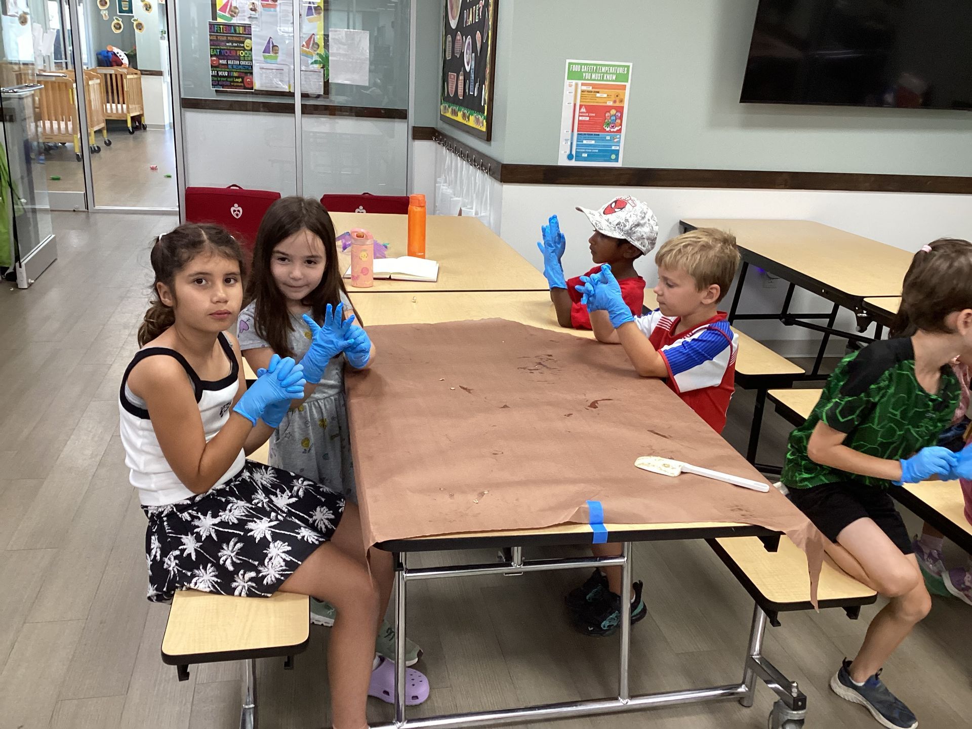 Children seated at a table in a classroom, wearing gloves and handling craft items