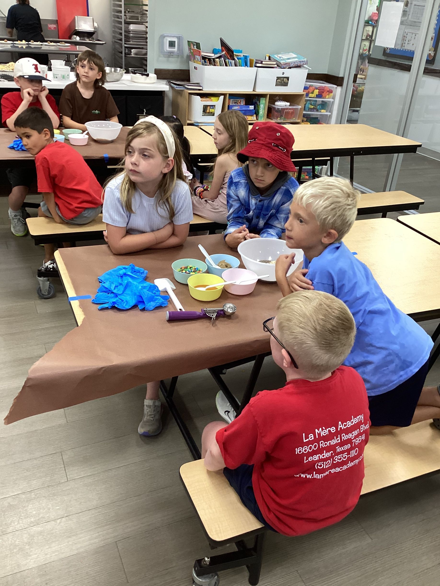 Children seated at a classroom table, eating snacks and talking during a group activity.