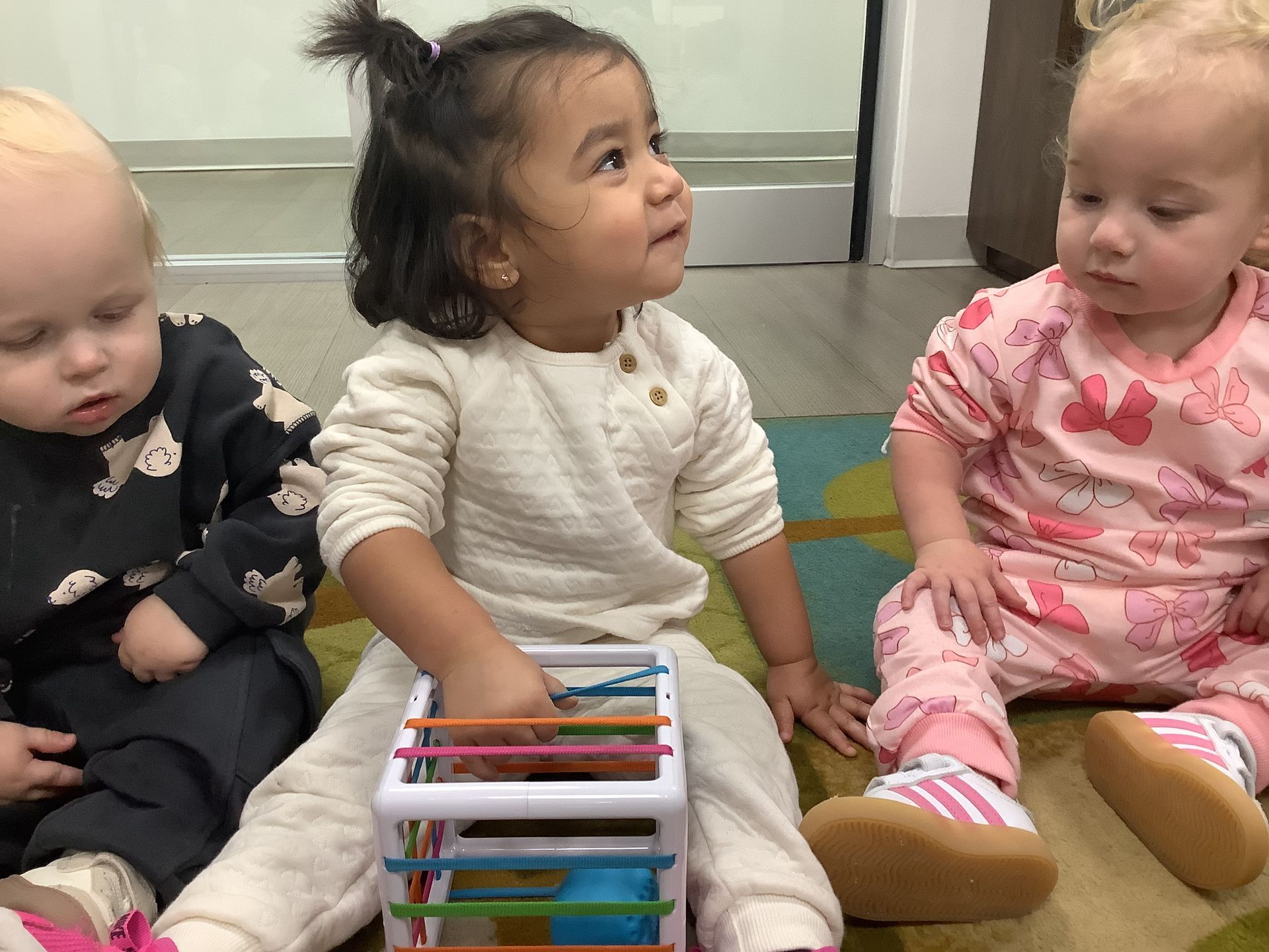 Three babies sitting on the floor with a colorful toy, one looking up, in a playroom