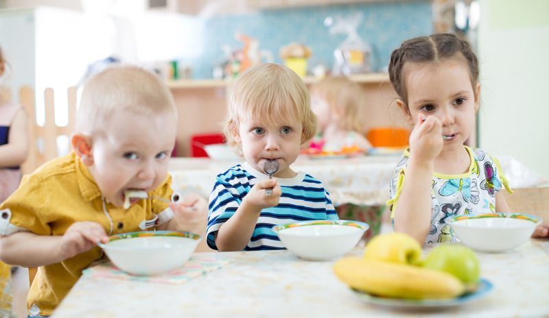 Three toddlers eating breakfast at a table with bowls and fruit in a bright kitchen.