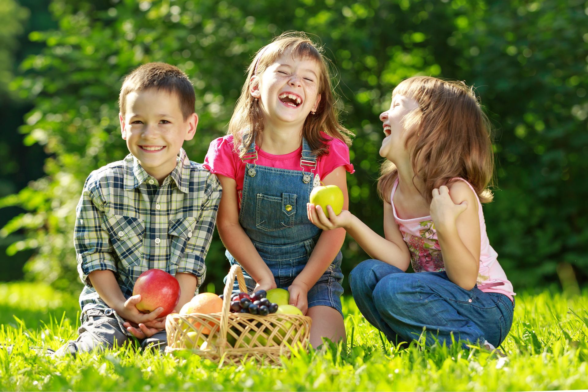 Three children sitting on grass with picnic baskets and fruit, smiling outdoors in a sunny park.