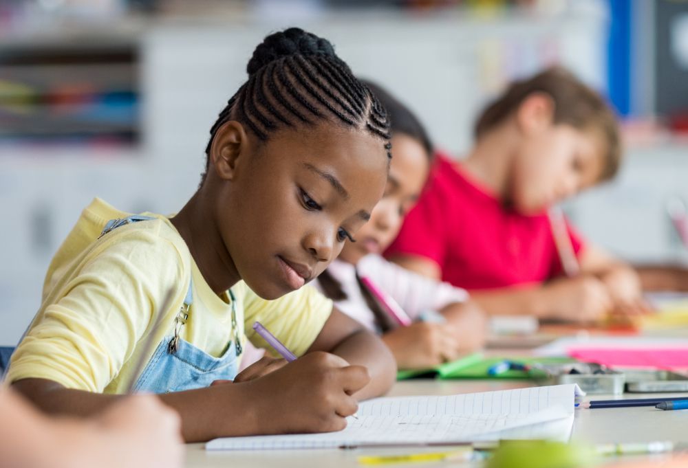 Students writing at desks in a classroom, with a child in a yellow shirt in the foreground.