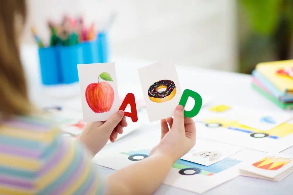 Child’s hands holding alphabet cards with fruit and donut illustrations on a bright desk