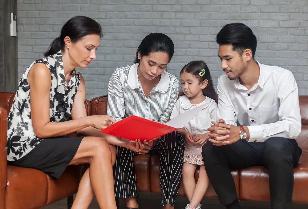 Four people sit on a couch looking at a red book together in a living room.