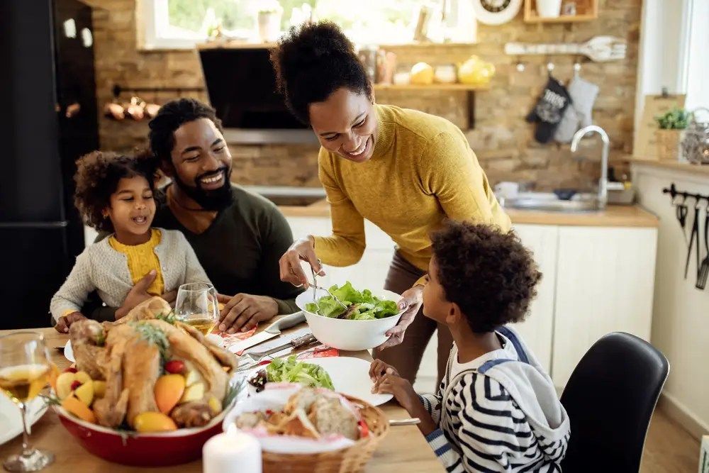 Family sharing a meal at a kitchen table, with smiling adults and children around a bowl of salad and dishes