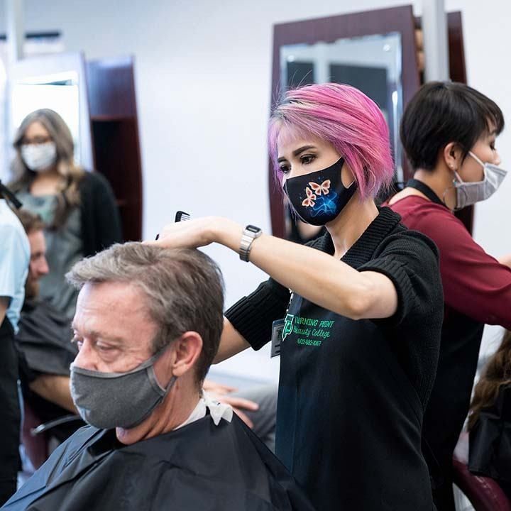 Hair stylist with pink hair, wearing a mask, cuts hair of a client at a salon.