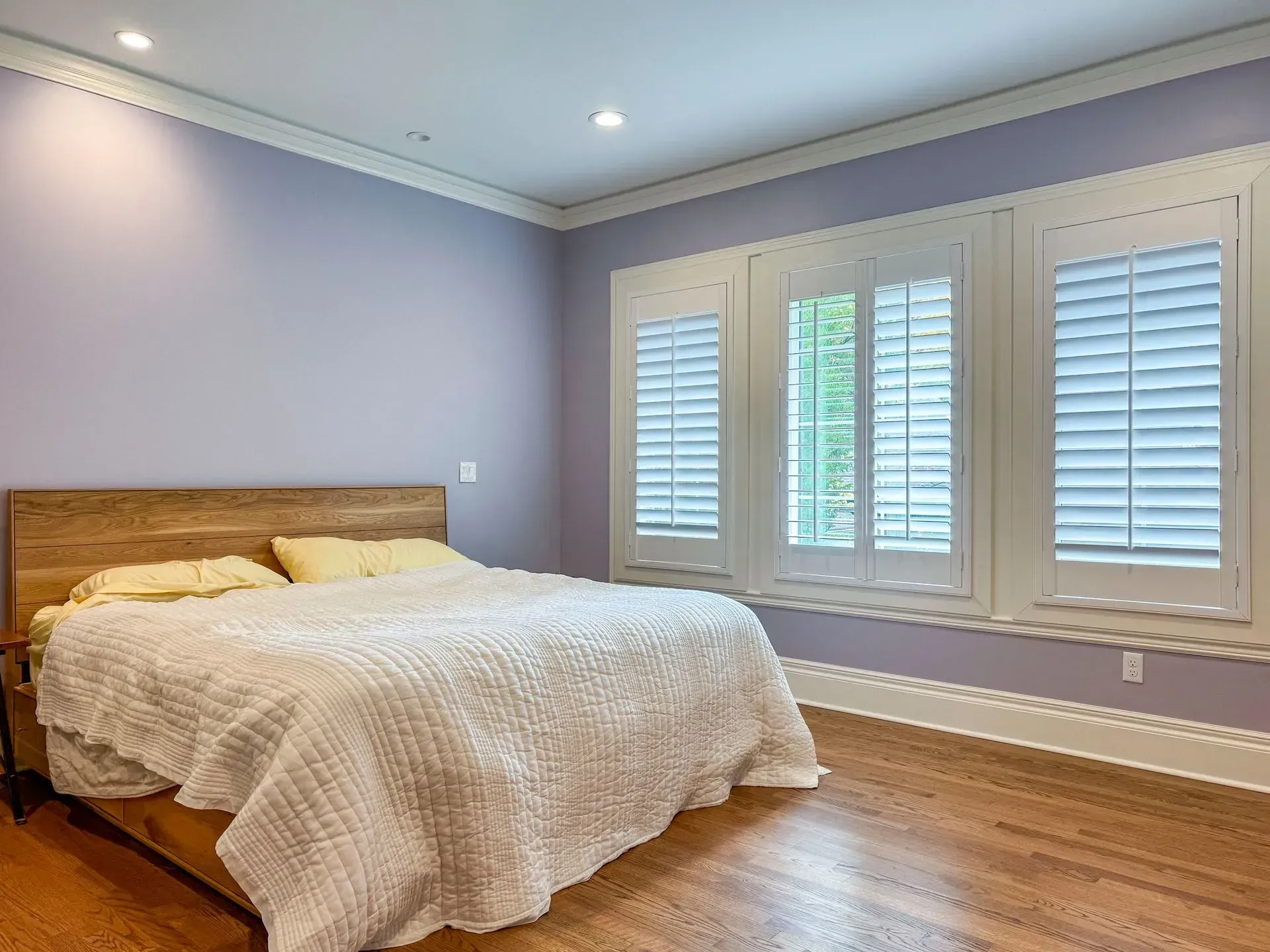 A bedroom with purple walls and white shutters on the windows.