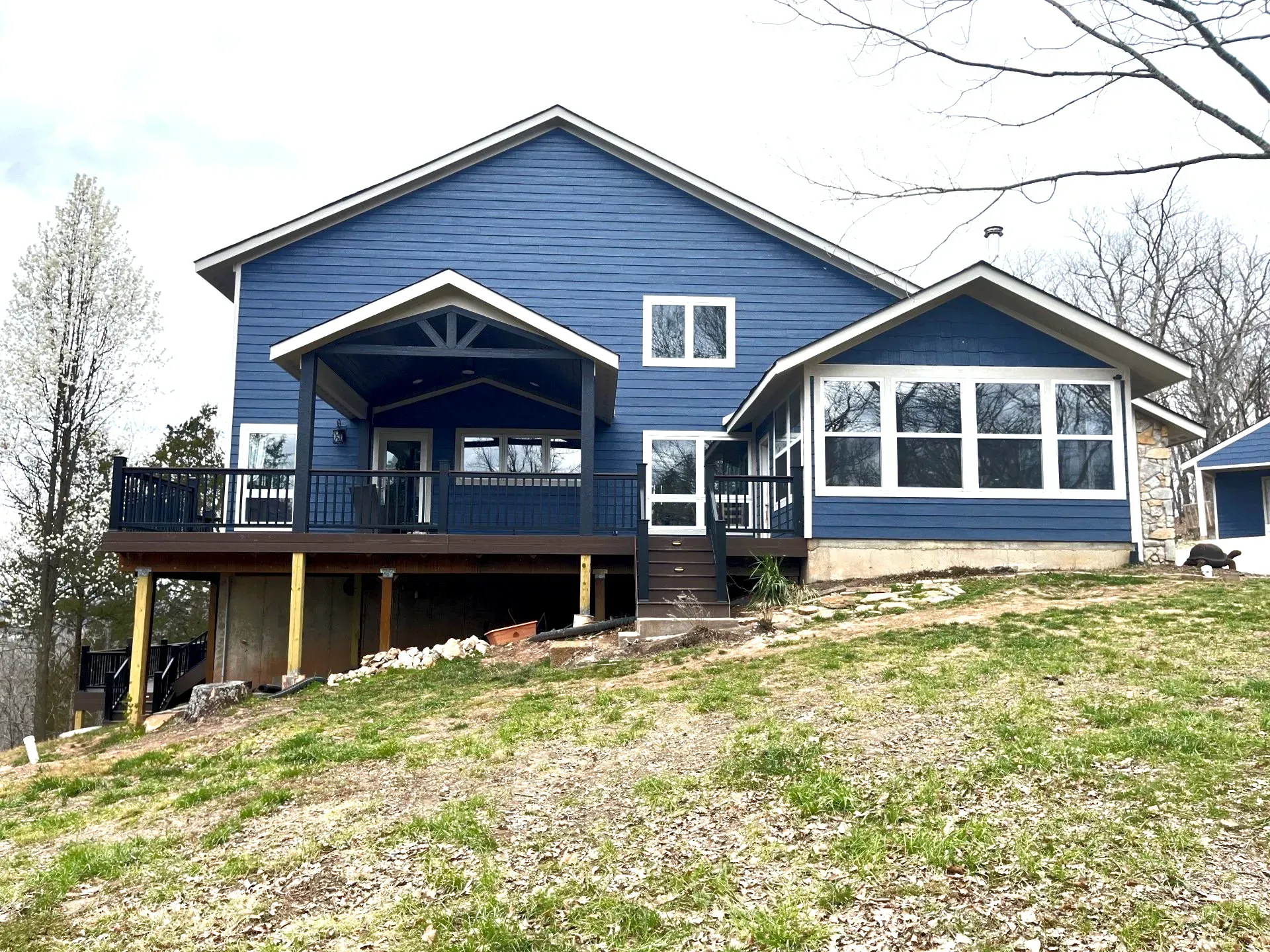 A large blue house with a large deck is sitting on top of a grassy hill.