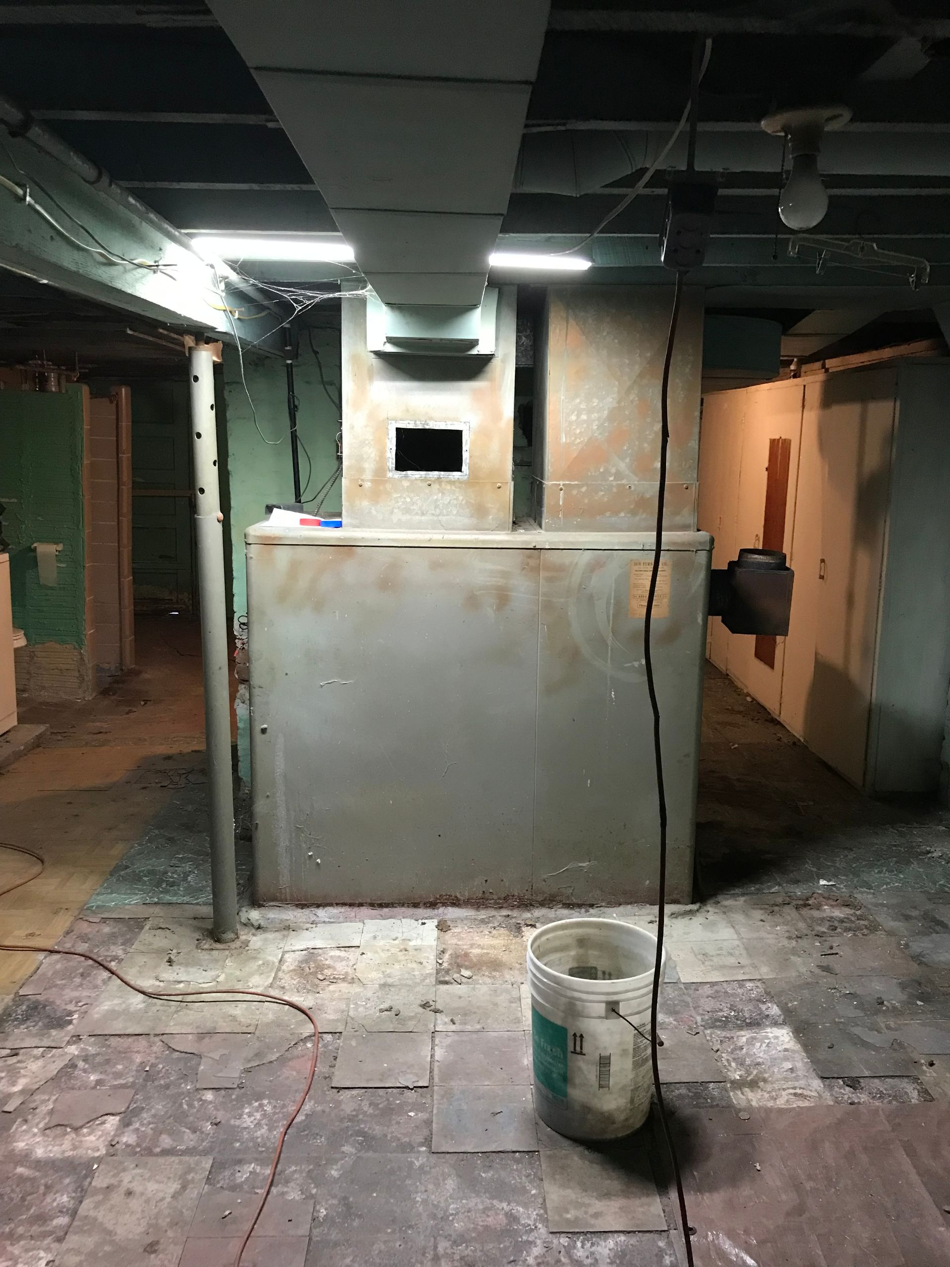 Old, dusty furnace in a basement setting. Metal exterior, ductwork overhead, brick floor, paint peeling.