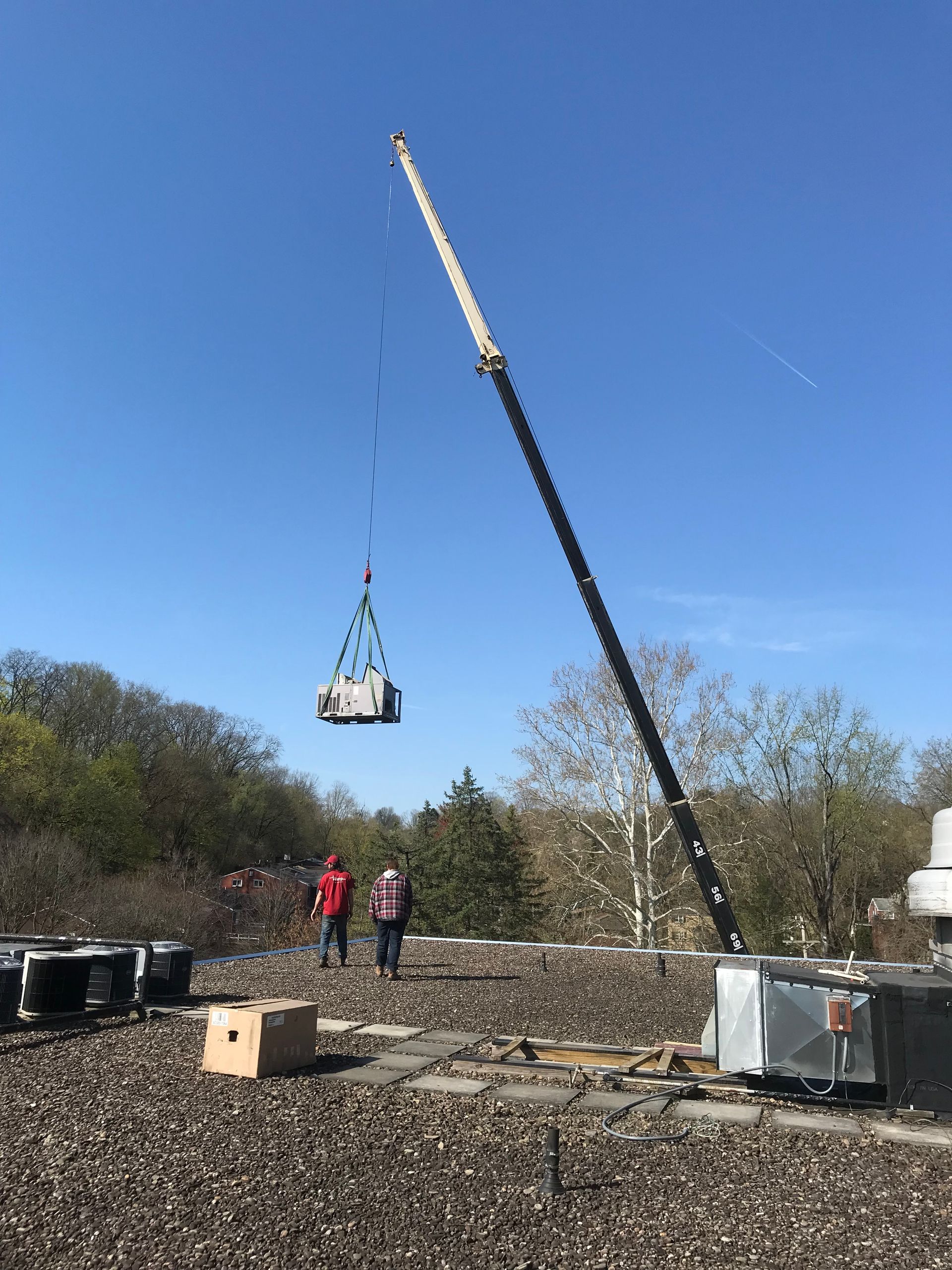 Crane lifting an HVAC unit onto a rooftop. Two workers observe. Blue sky and trees in the background.