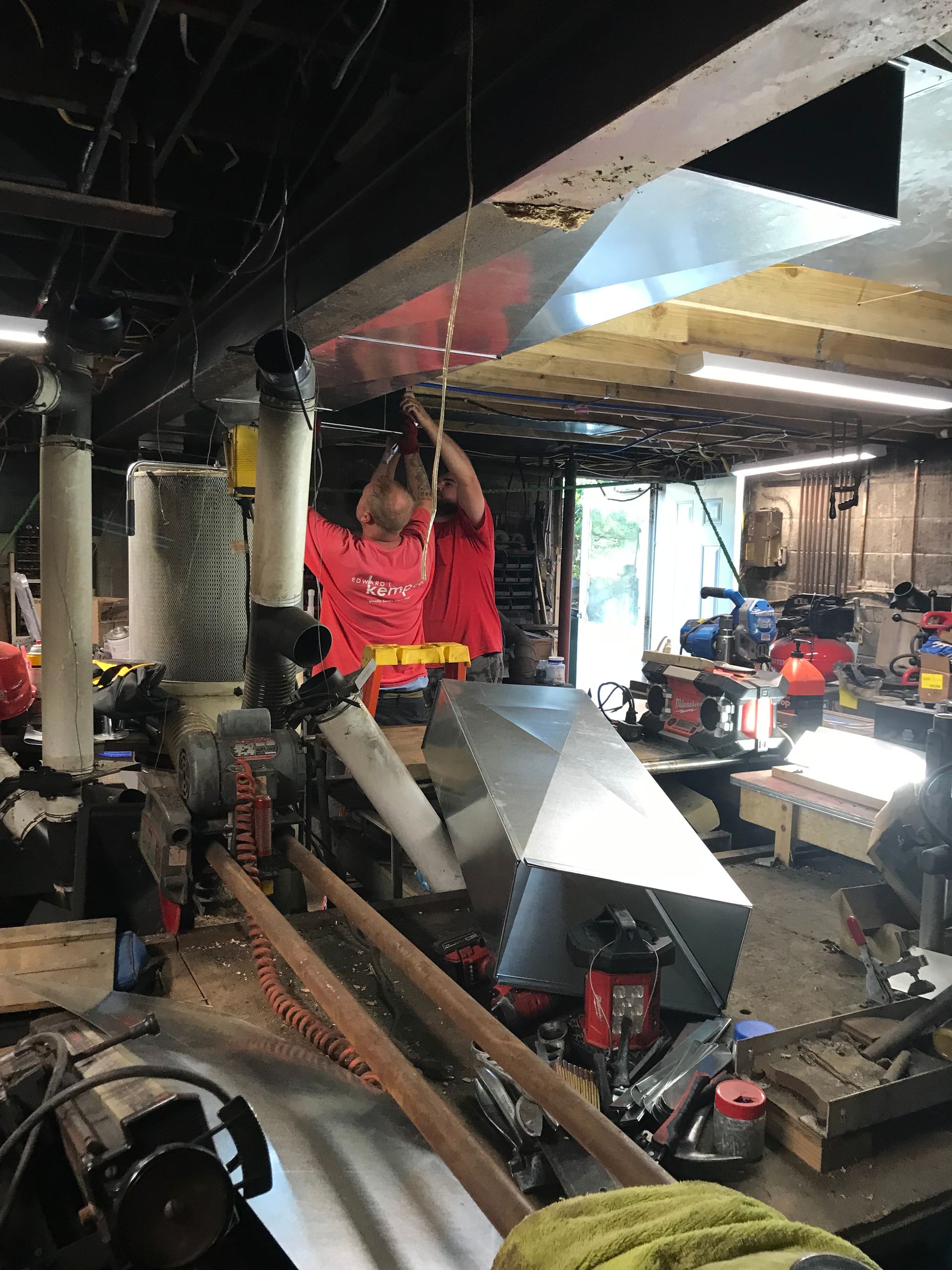 Two people installing metal ventilation ductwork in a cluttered workshop.
