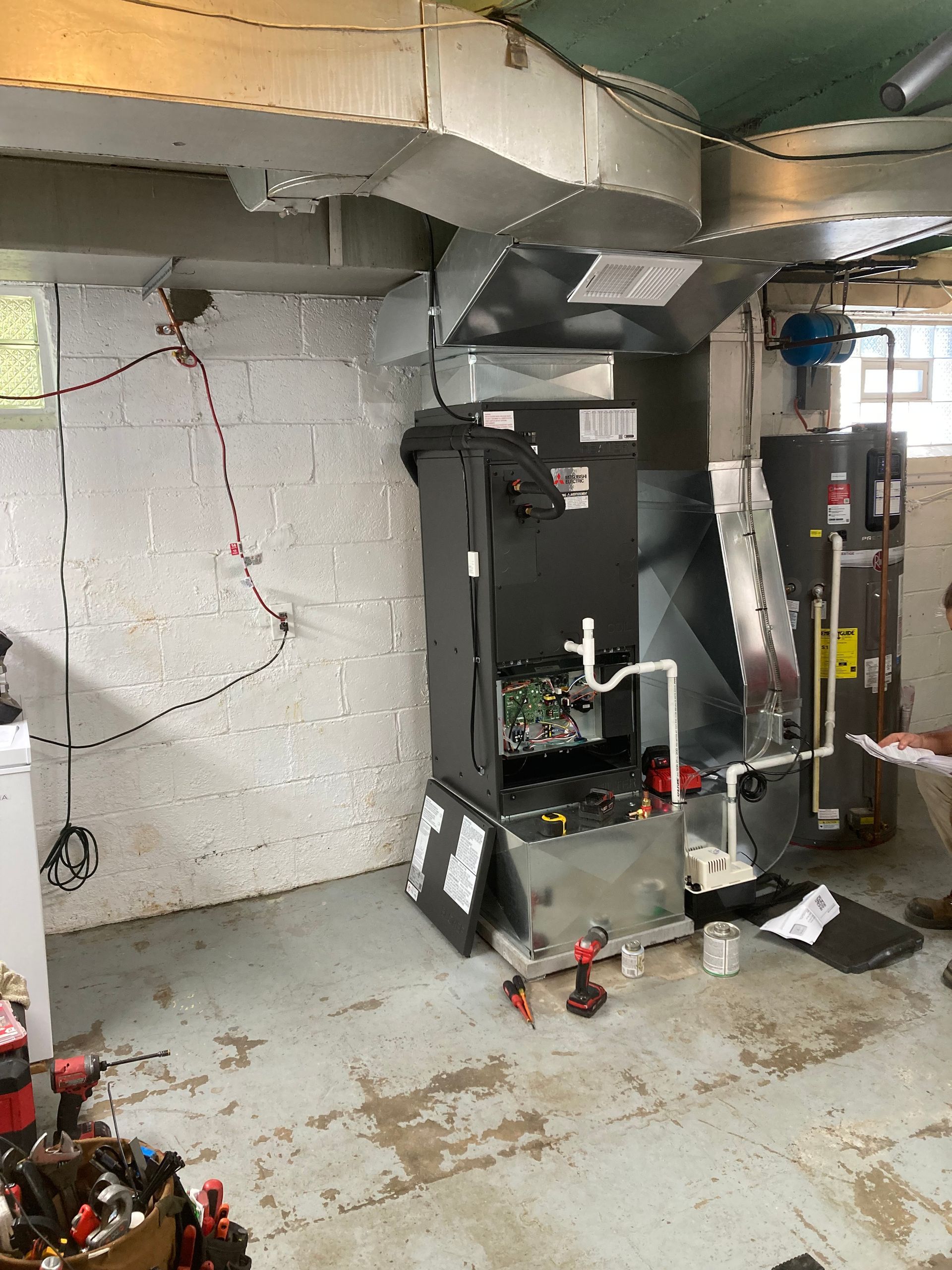Furnace installation in a basement; silver ductwork, water heater, tools, concrete walls.