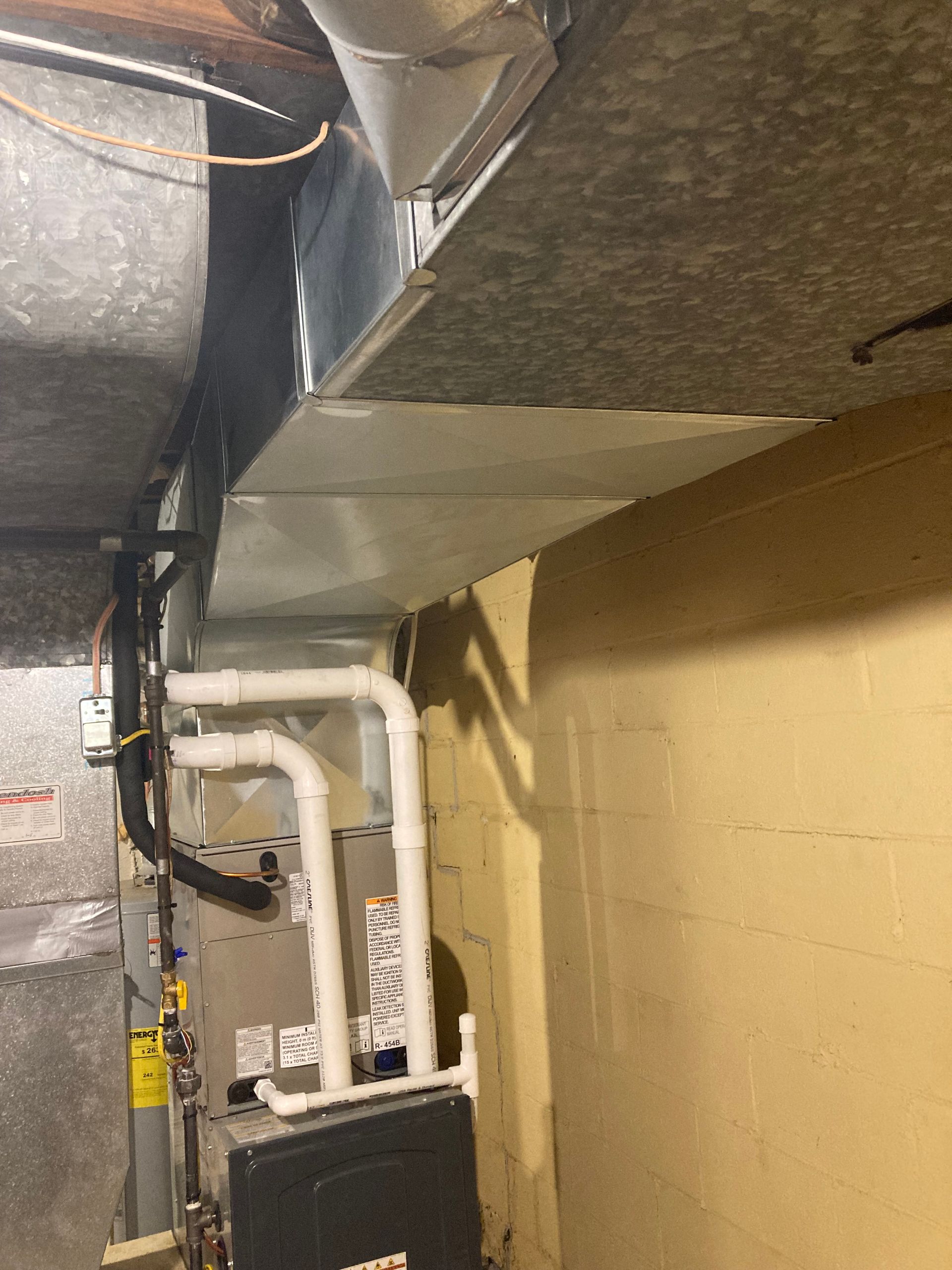 Furnace and ductwork in a basement setting; metal and white pipes against a yellow wall.