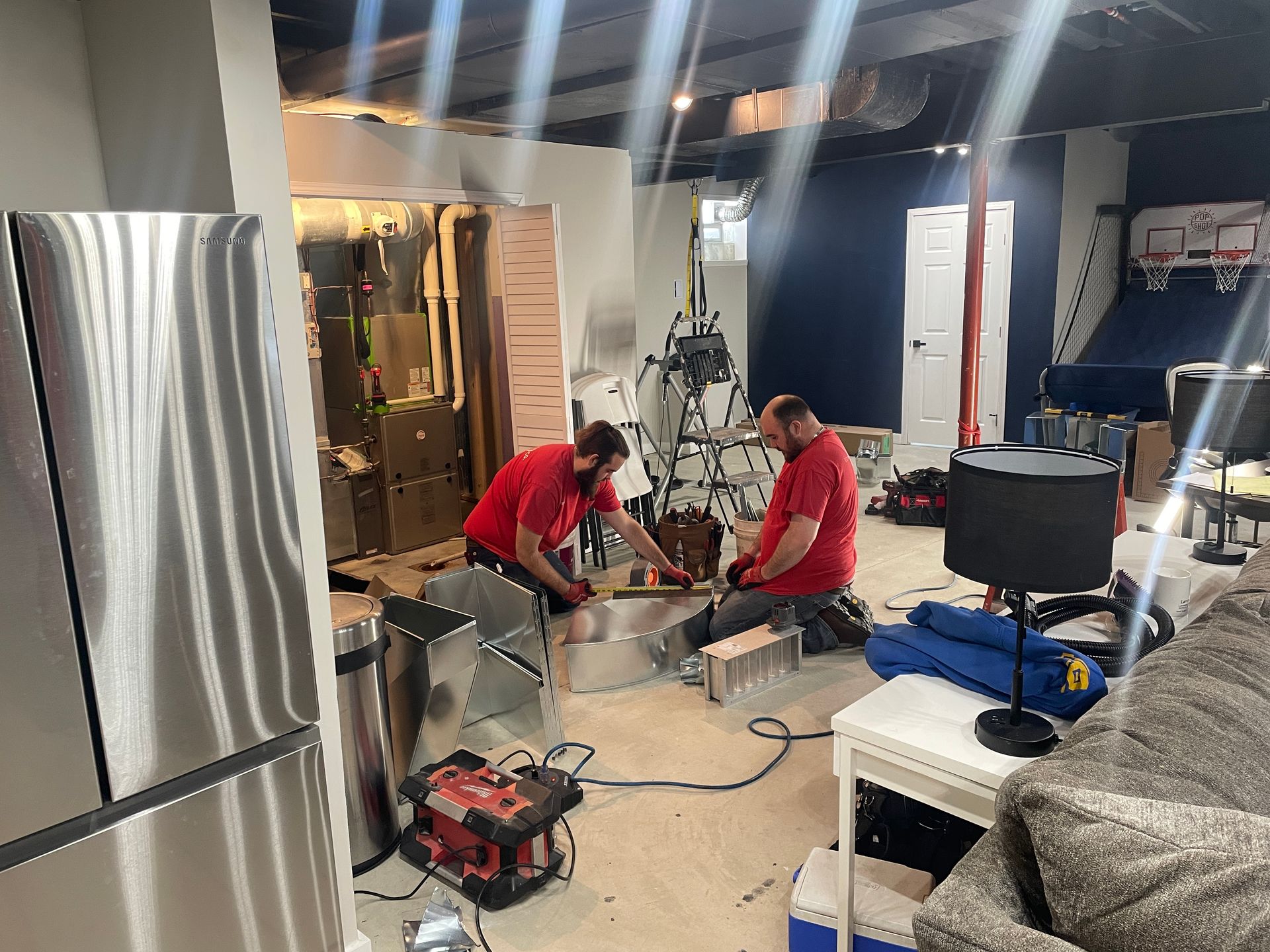 Two men in red shirts working on a project in a basement, next to a refrigerator, with tools.