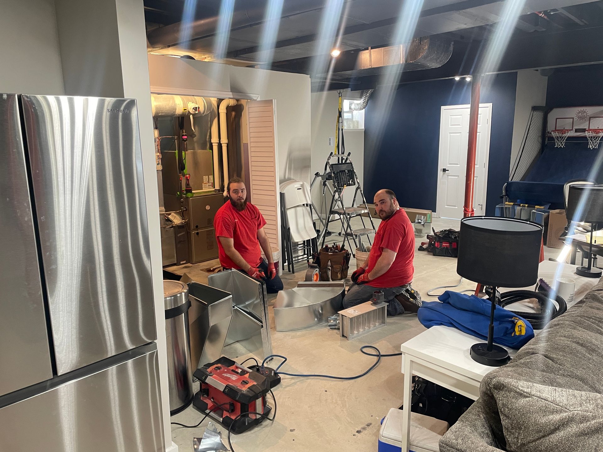 Two men in red shirts working on a basement renovation, with tools, appliances, and exposed walls.