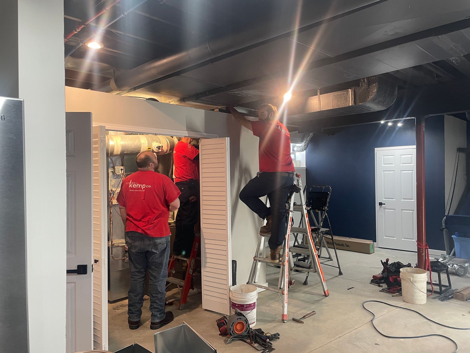 Three workers in red shirts installing panels inside a room. Ladders, tools, and a white door are visible.