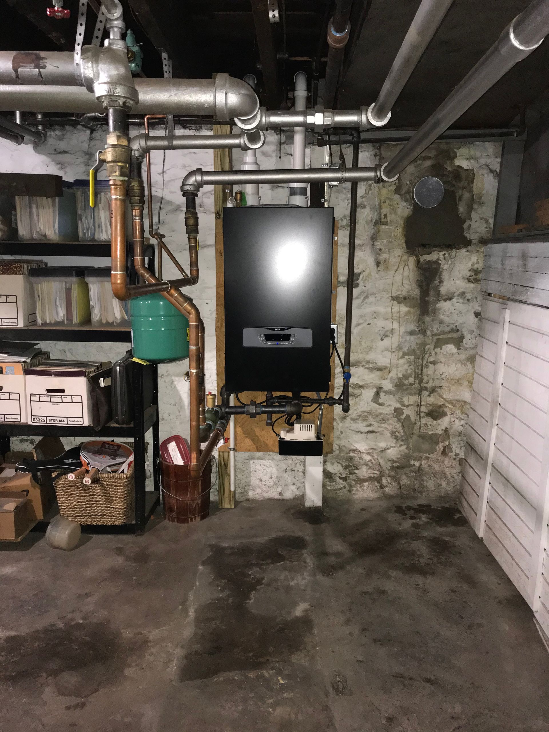 Black heating unit and associated copper pipes against a concrete wall in a basement setting.