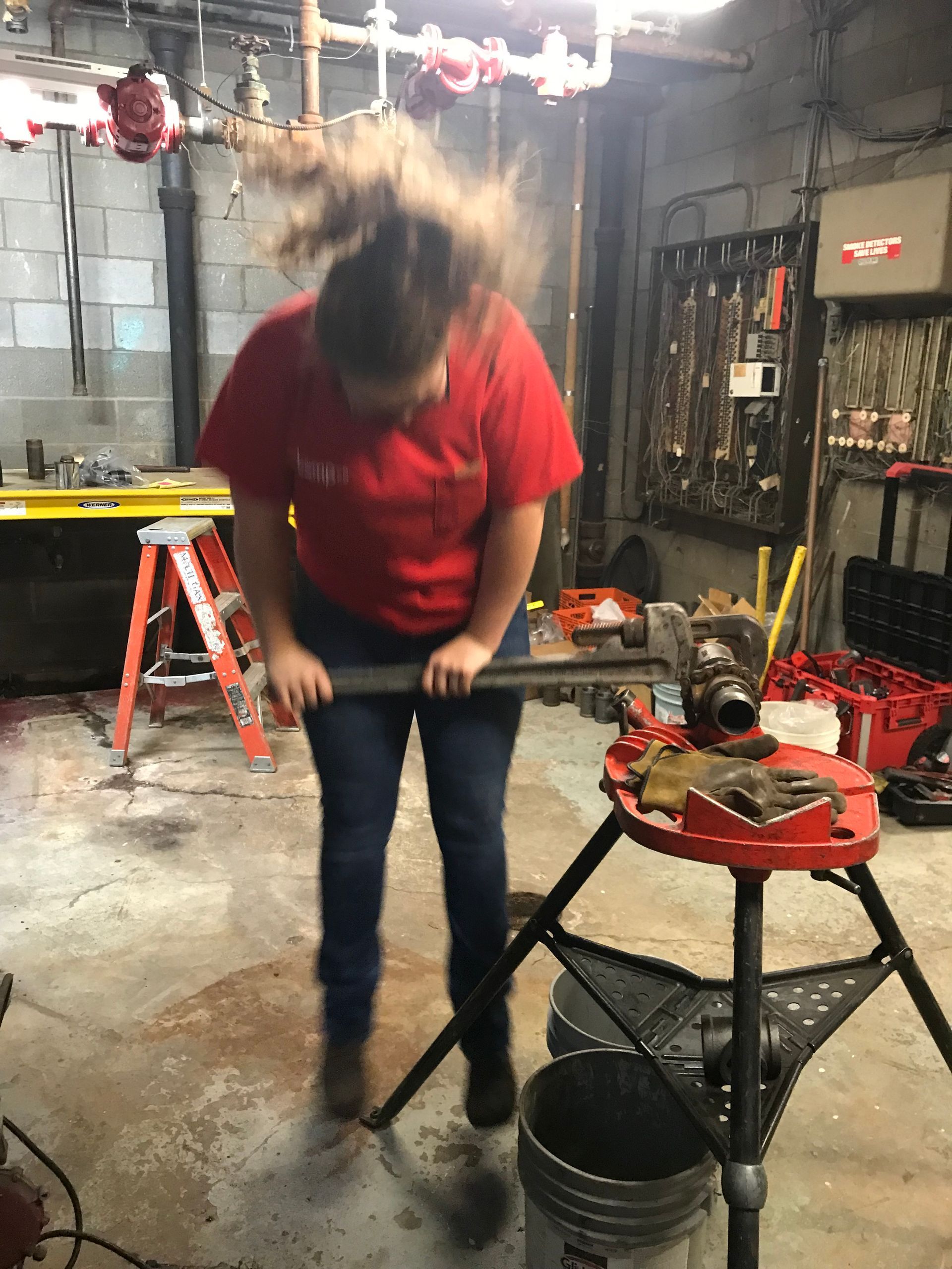 Person in red shirt and jeans, straining to tighten a wrench in a cluttered workshop.