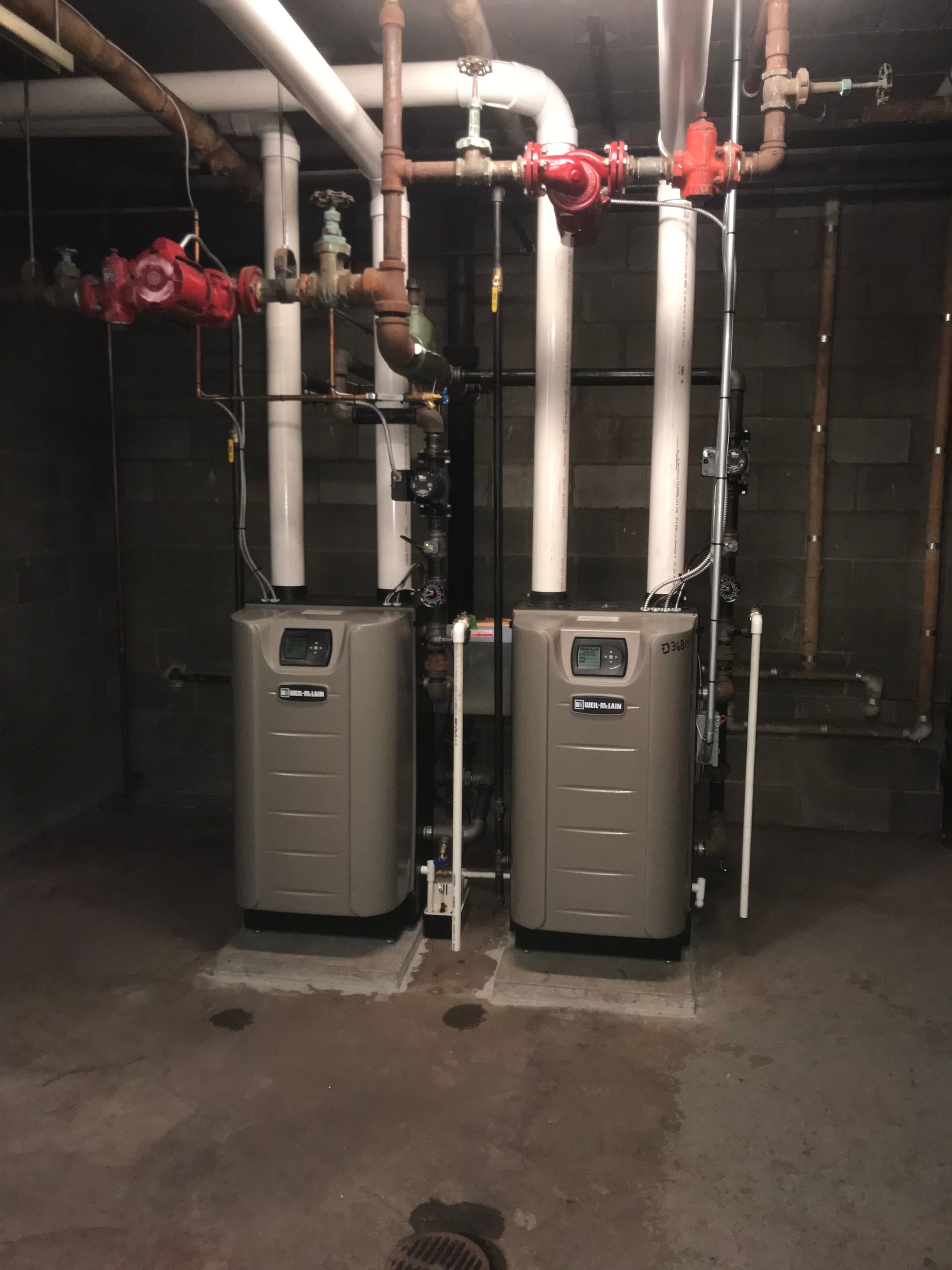 Two tan heating units with white insulated pipes and red valves in a basement setting.