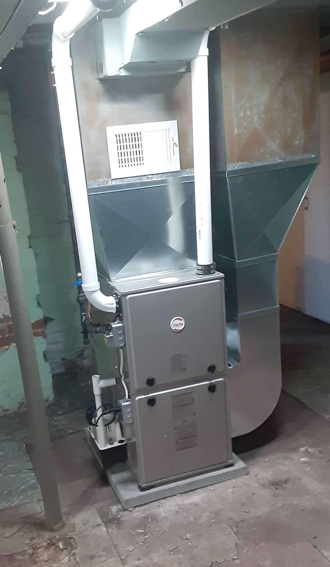 Furnace unit in a basement, with silver ductwork, white pipes, and a gray metal casing.