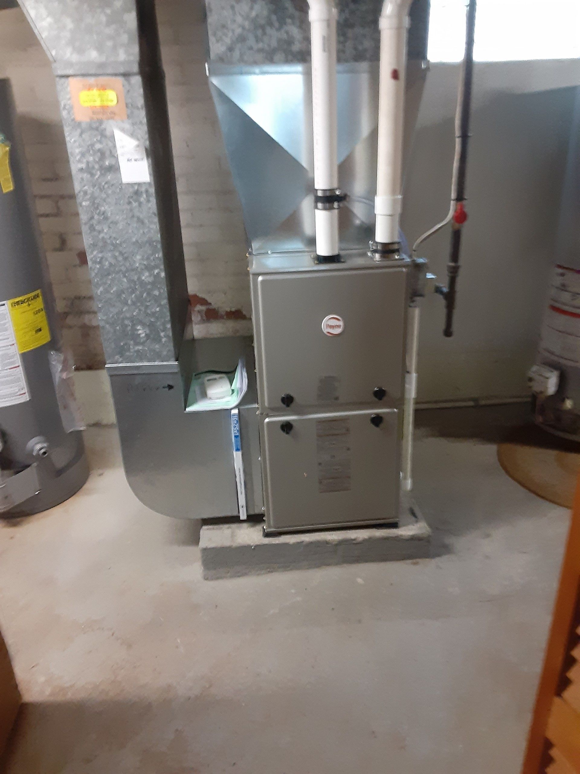 Furnace and ductwork in a basement. Grey metal unit with white pipes and air vent. Concrete floor.