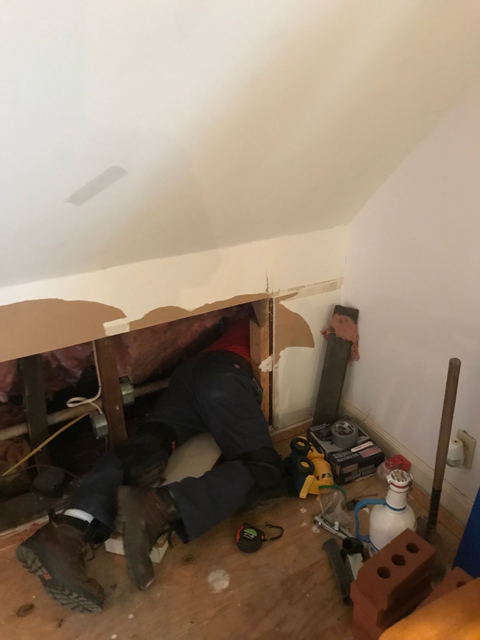 Person working on plumbing, crouched in a small room. Tools and materials are scattered on the floor.