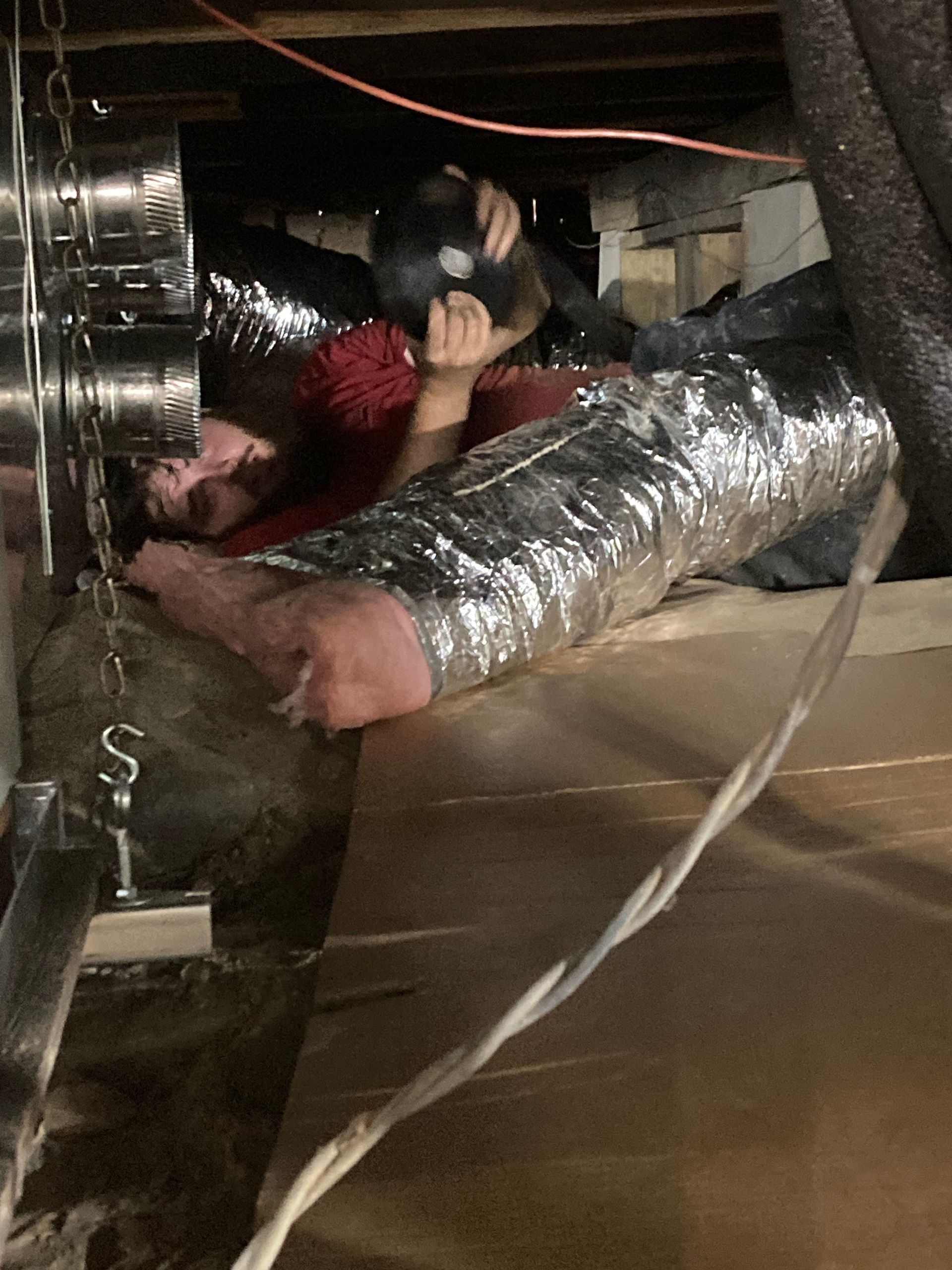 Person lying in a crawl space, working near a silver flexible duct and electrical wires.
