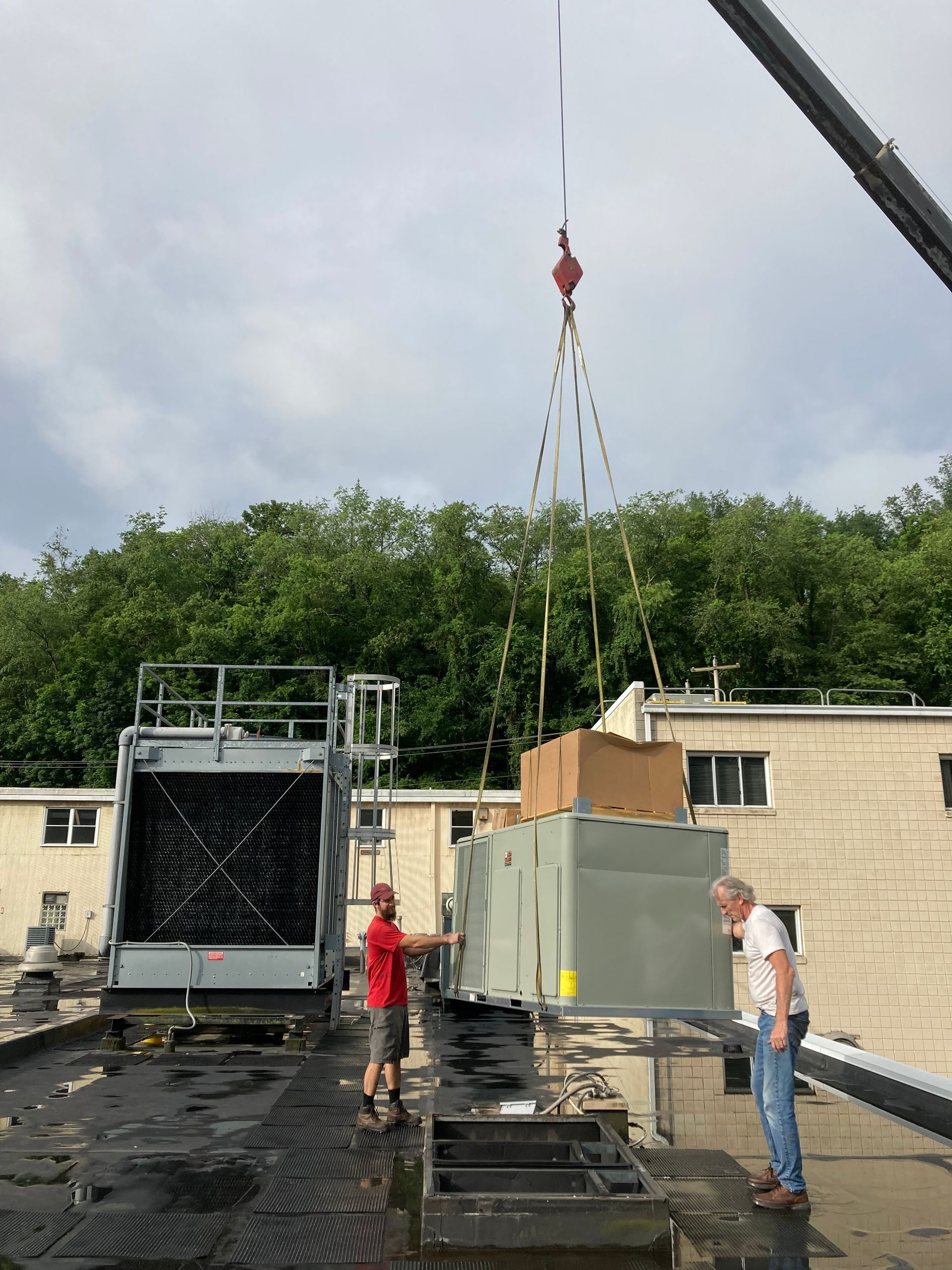 Crane lifting HVAC unit onto a rooftop. Two men observe near an existing unit and trees in the background.