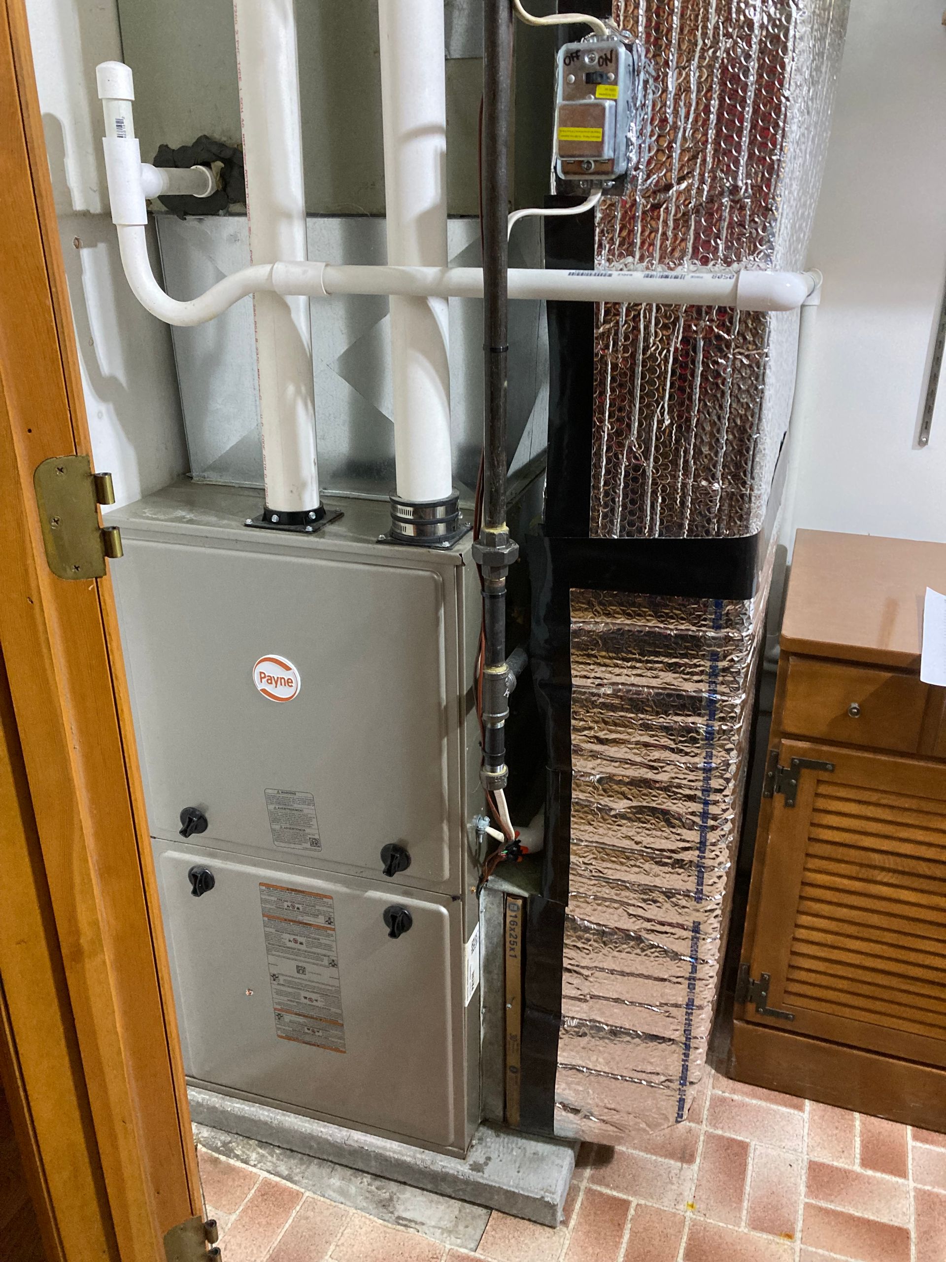 Furnace unit in a basement. Grey metal box, with white pipes, next to air conditioning coils.