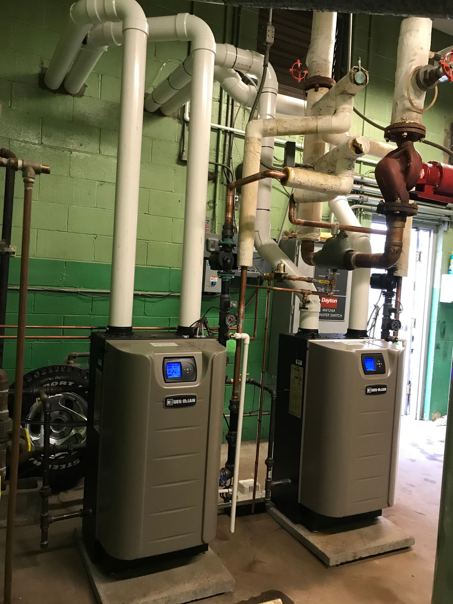 Two gray boilers with white pipes in a green-walled utility room.