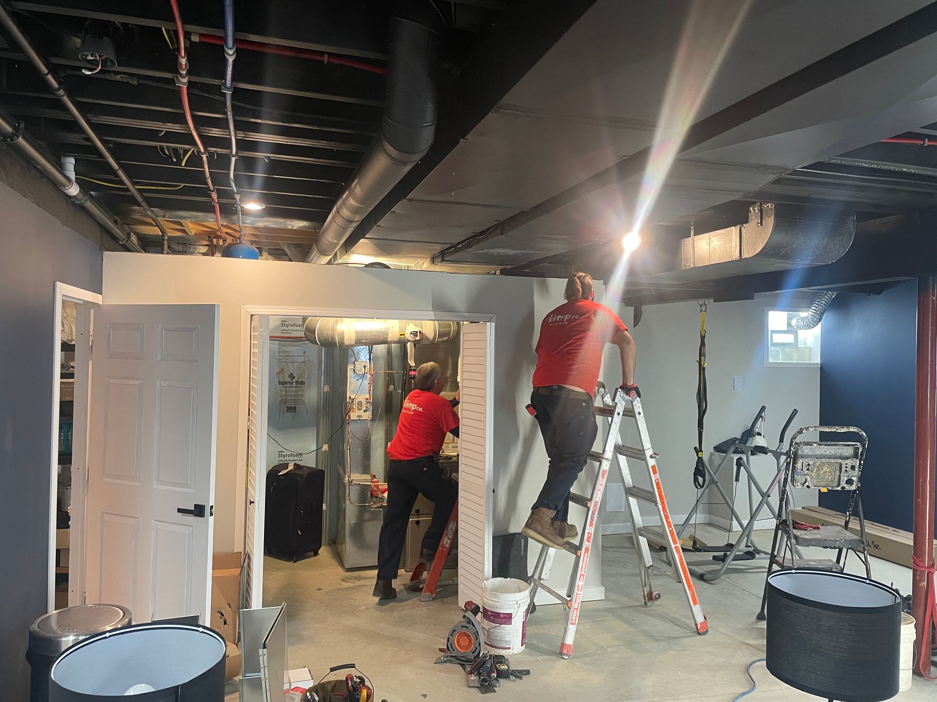 Two people in red shirts are working in a basement; one on a ladder, the other near a furnace.
