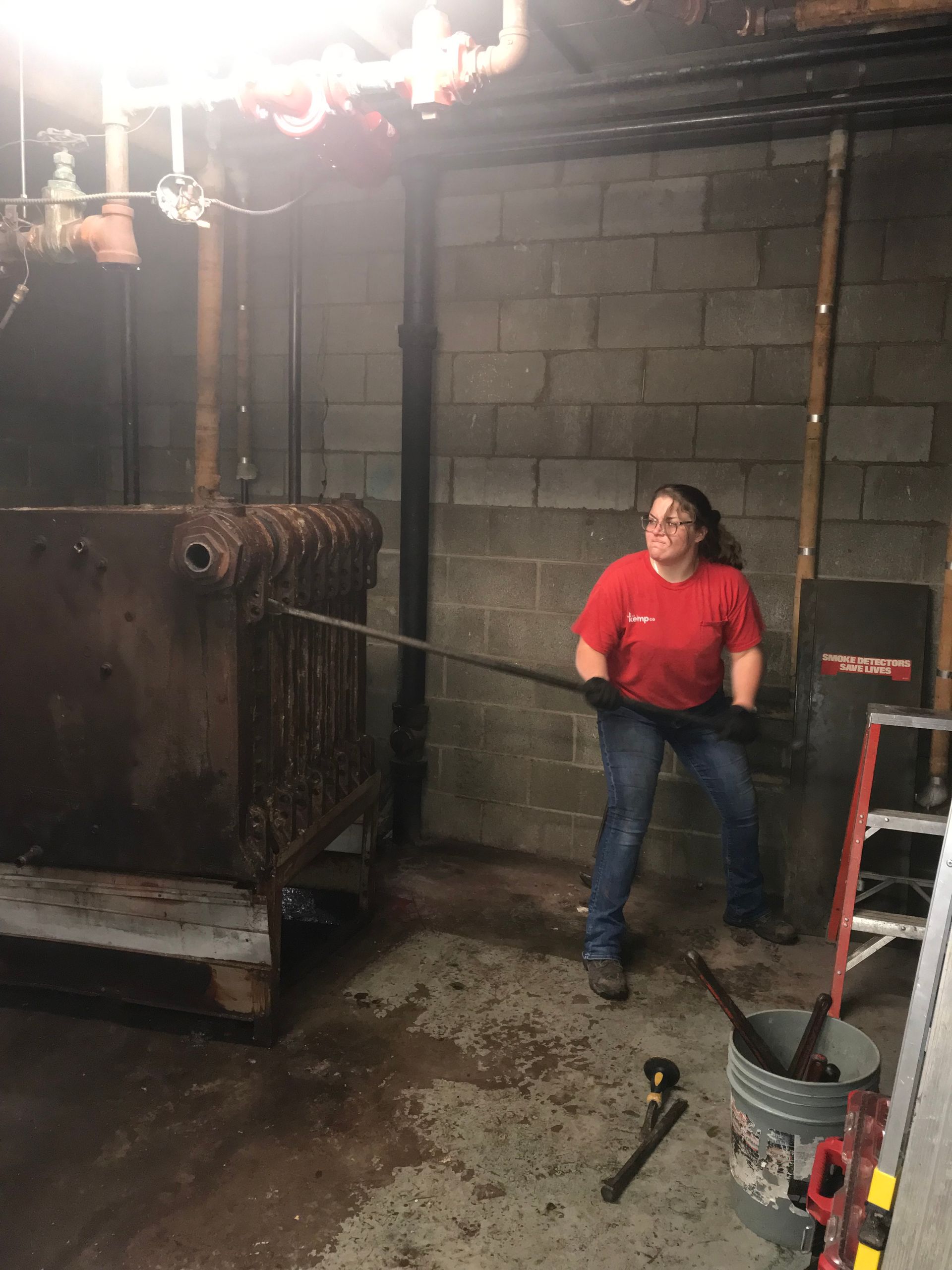 Woman in red shirt and jeans uses a tool to work on a large rusty radiator in a basement setting.