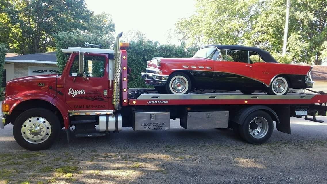 Red tow truck hauling a red classic car on a flatbed in a driveway.