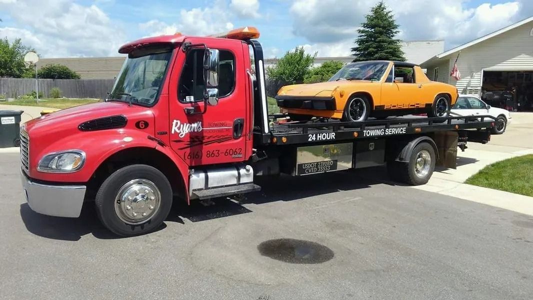 Red flatbed tow truck carrying an orange pickup on a residential street under a blue sky