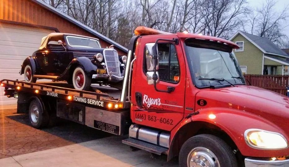 Red tow truck carrying a small black SUV on a flatbed in a residential driveway at dusk