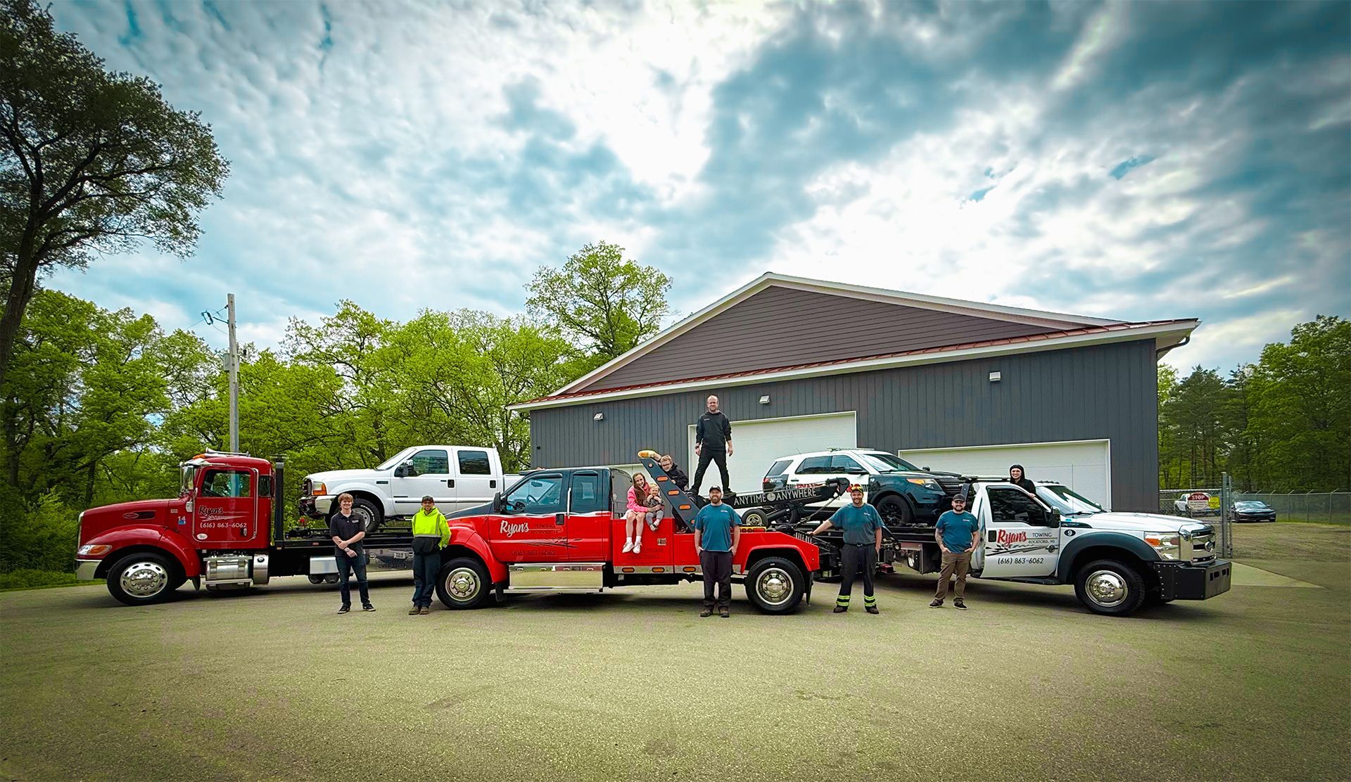Tow truck scene with several pickup trucks and people lined up outside a large garage under a cloudy sky