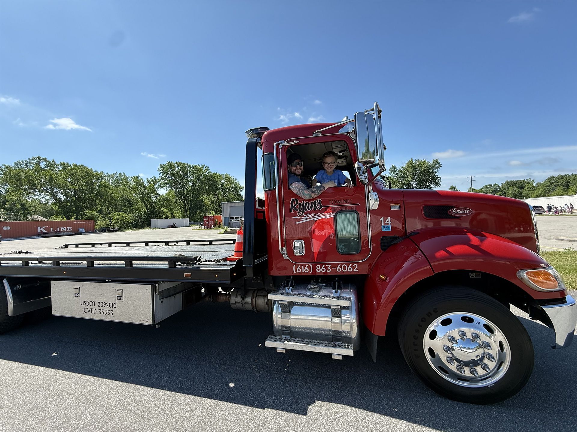 Red flatbed tow truck with driver on a sunny road, trees and blue sky in the background