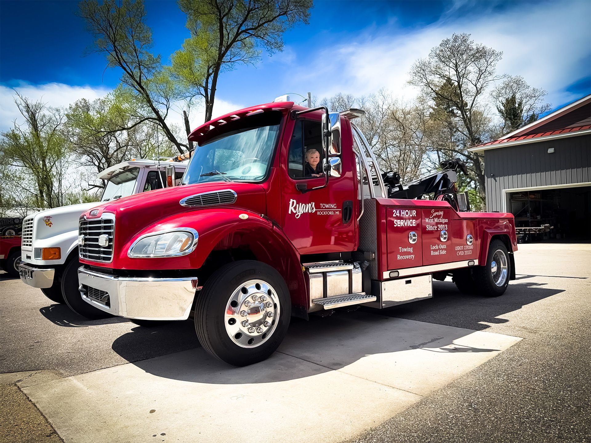 Red tow truck parked outdoors beside a building, with trees and blue sky in the background.