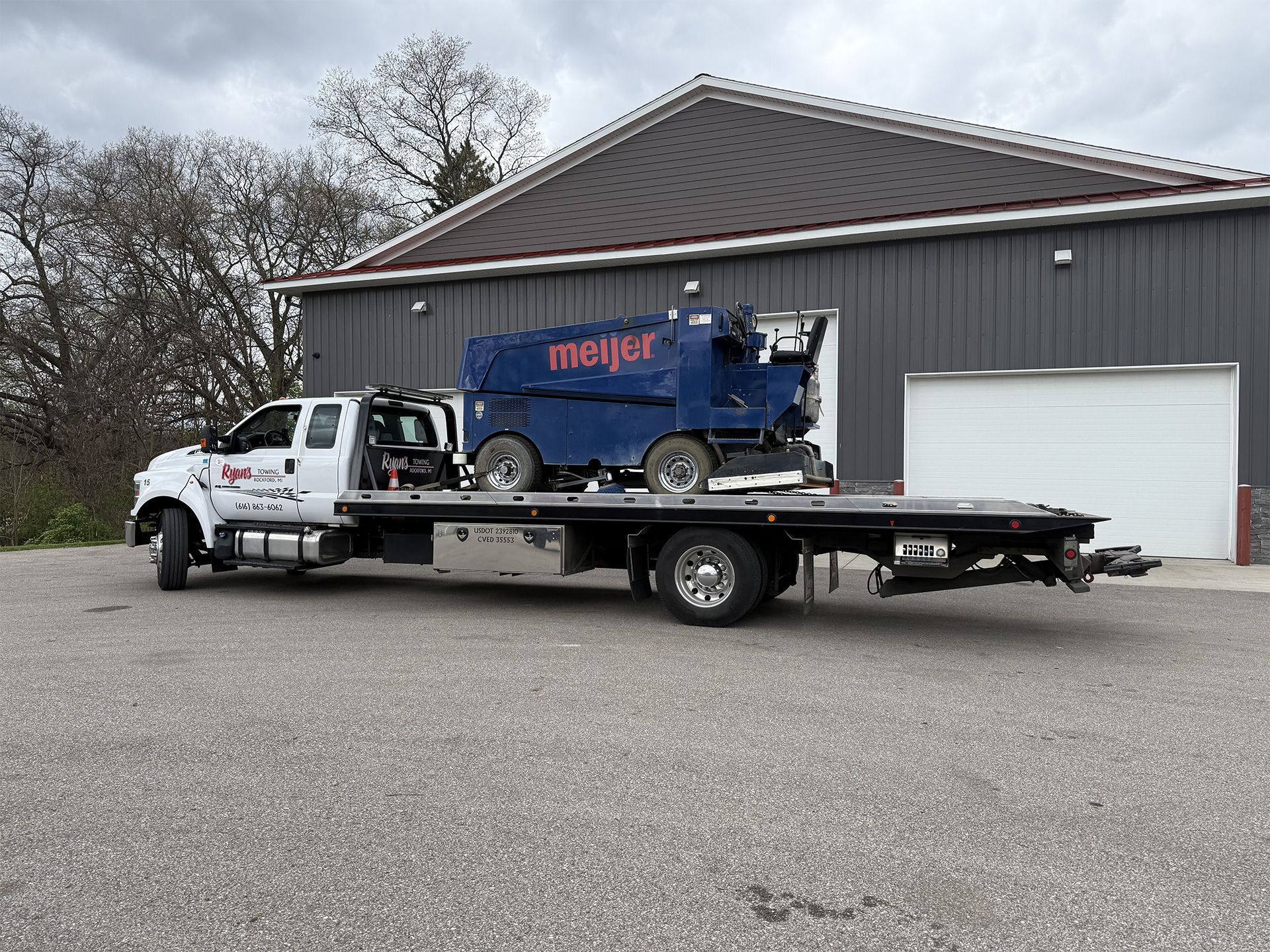 Tow truck hauling a blue Meijer dumpster trailer in front of a gray industrial building