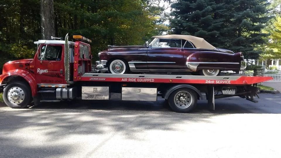 Red tow truck carrying a maroon classic car on a flatbed in a tree-lined street.