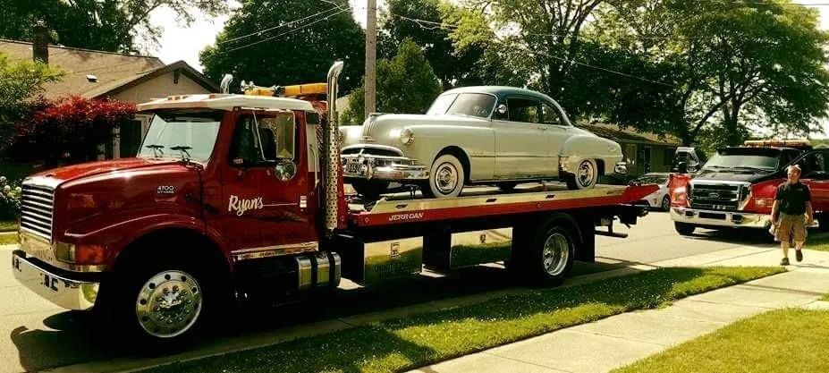 Red tow truck hauling a silver classic car in a suburban neighborhood during daylight.