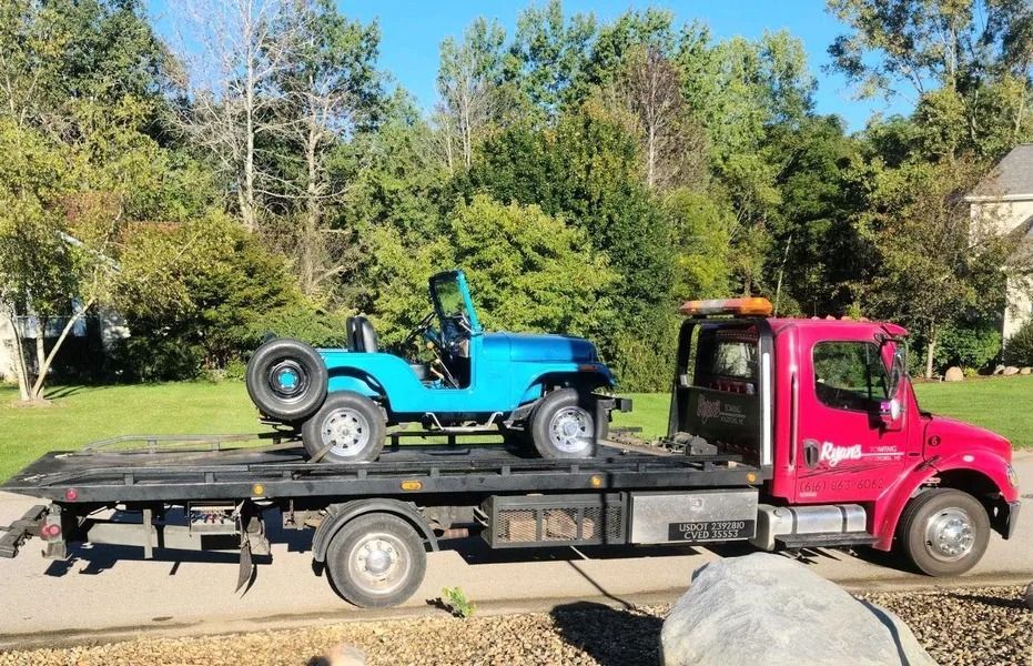 Pink tow truck carrying a blue Jeep on a flatbed trailer beside trees and grass