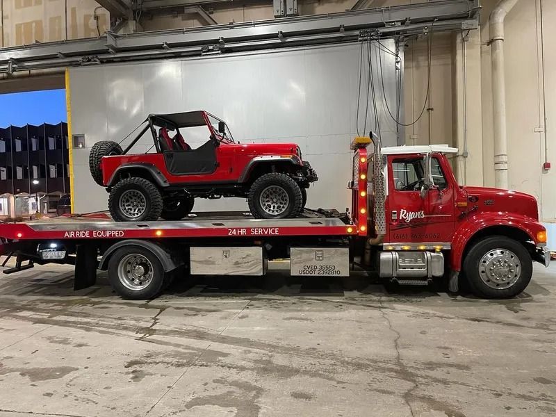 Red tow truck carrying a red off-road Jeep in an indoor garage.
