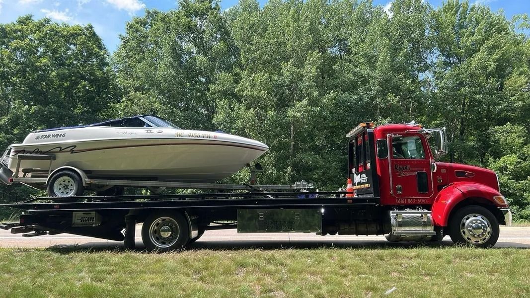 Red tow truck hauling a white speedboat on a trailer by a grassy roadside with trees behind