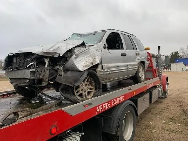 Severely damaged silver sedan loaded on a red tow trailer outdoors under cloudy sky