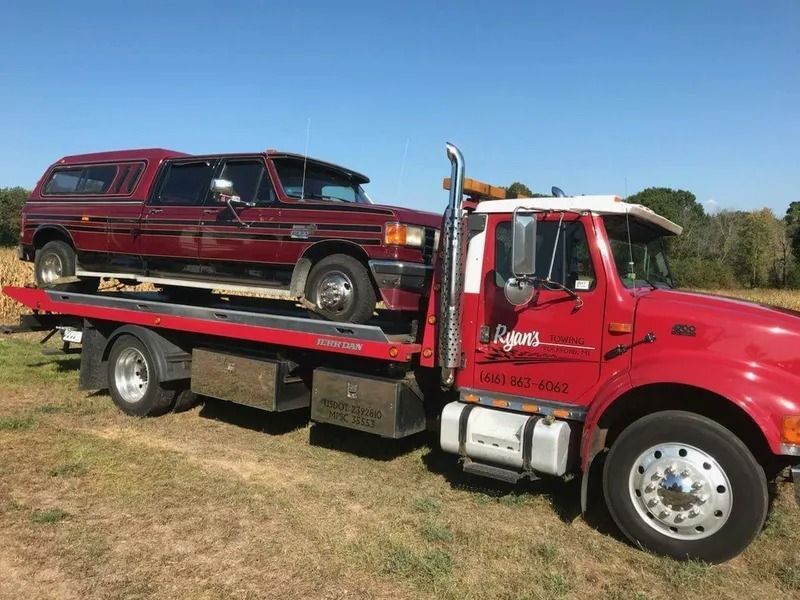 Red flatbed tow truck hauling a maroon pickup on a grassy field under a clear blue sky
