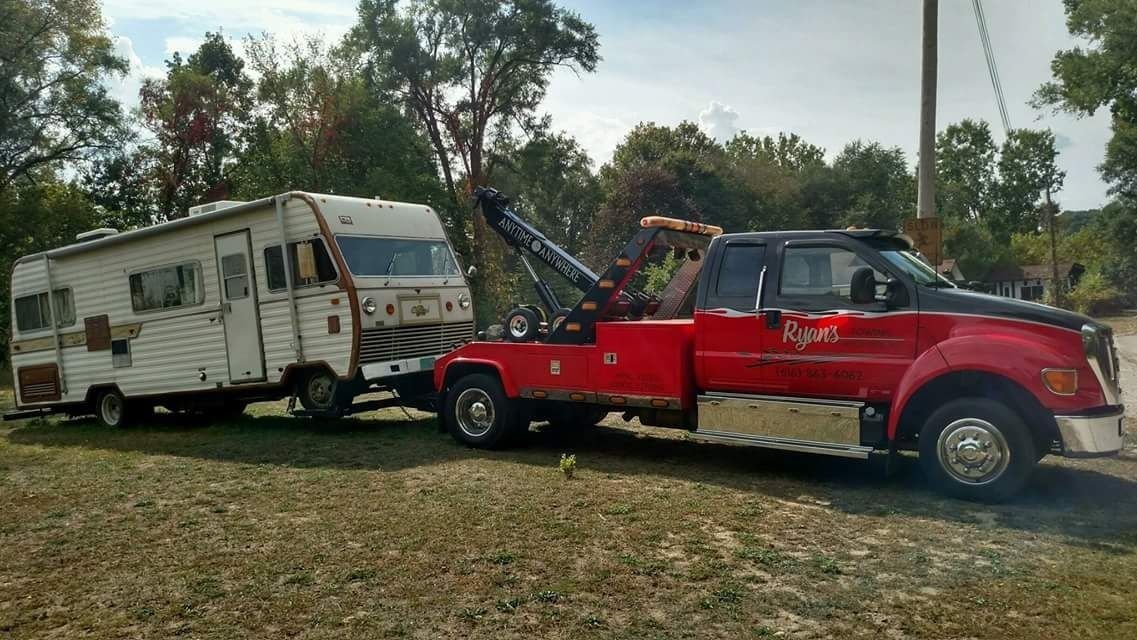 Red tow truck pulling a white camper trailer on a grassy roadside with trees in the background