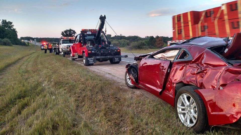 Red car badly wrecked in a field beside tow trucks and a highway at sunset