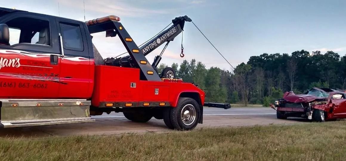 Red tow truck lifting a damaged maroon car in a grassy field near trees.