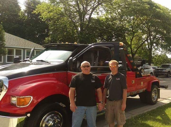 Two men standing beside a red and black tow truck outdoors in a wooded yard